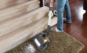 Photo shows legs and feet of man vacuuming throw rug at the foot of carpeted stairs.