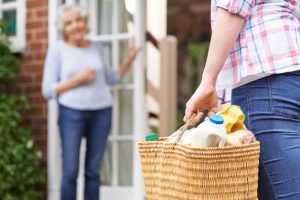 Person delivering groceries in a basket to older woman standing at doorway of her home.