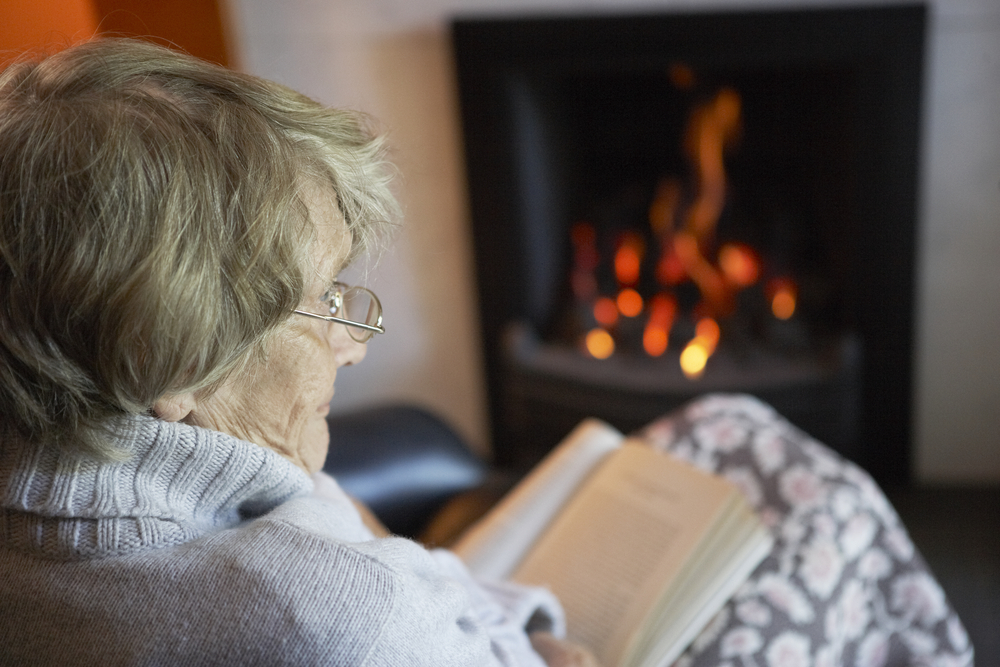 Older woman reading book by a fireplace.