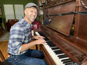 Jeffery Straker, musician, sits at older upright piano. He is wearing blue jeans, a blue plaid shirt, and a gray newsboy-style cap. He is smiling. A boom microphone is near him.
