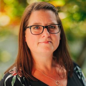 Thumbnail headshot of Bobbie Symes. Woman with long brown hair, floral top, against a blurred outdoor background