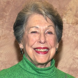 Lyn Goldman headshot. Smiling woman with styled grey hair wearing green turtleneck